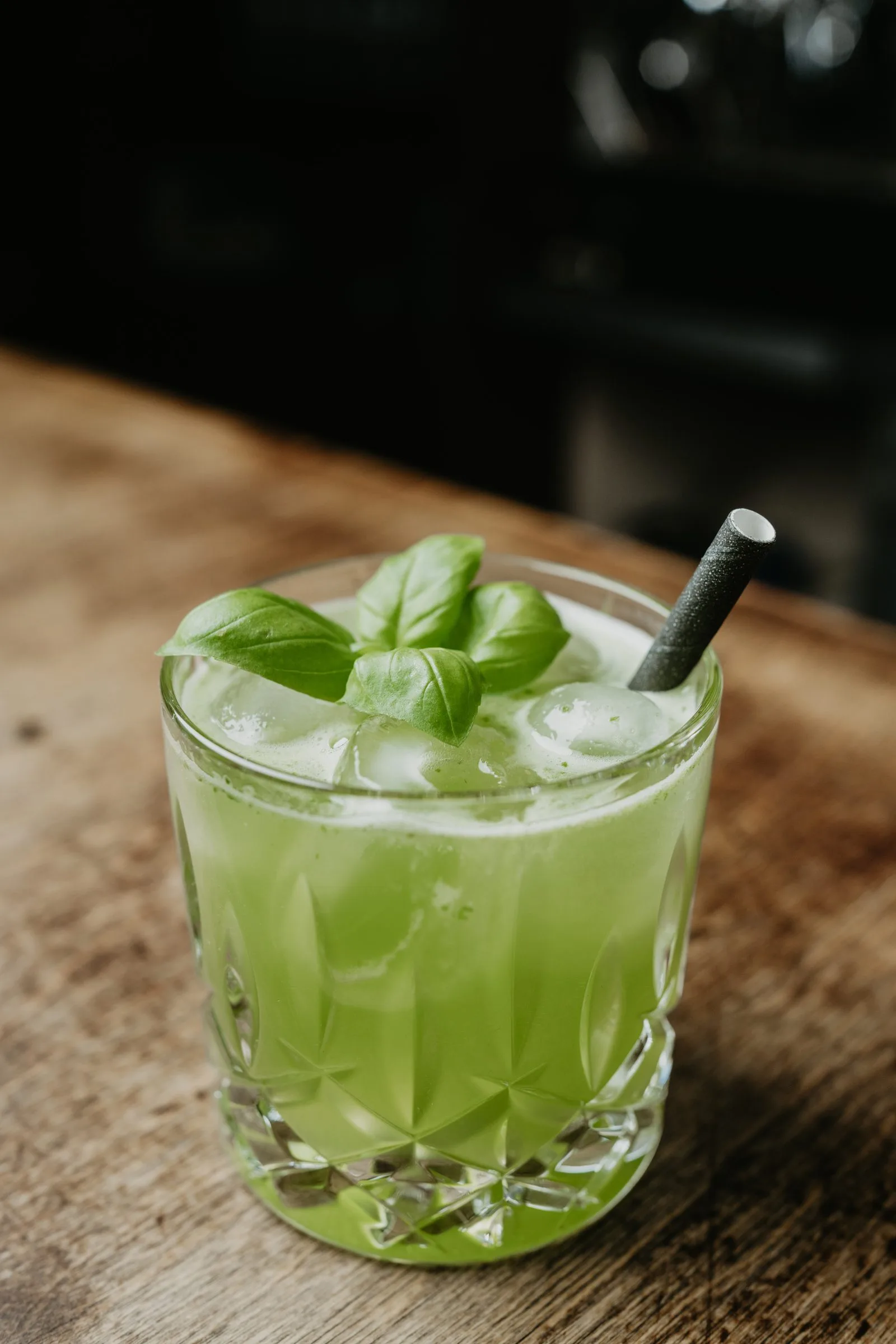 Ein grüner Cocktail mit Basilikum und Strohhalm im Kristallglas auf dem Holztresen.