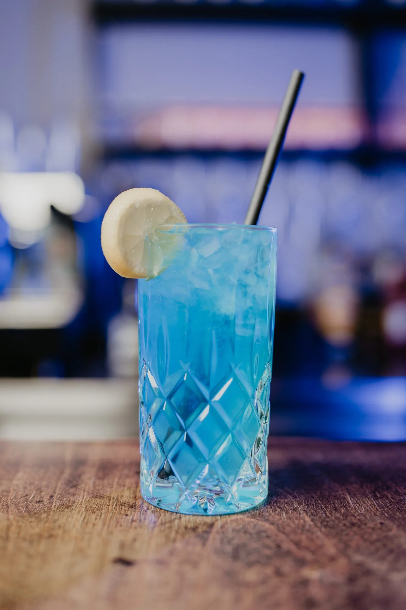Ein blauer Longdrink mit Zitronenscheibe vor blau beleuchtetem Hintergrund.