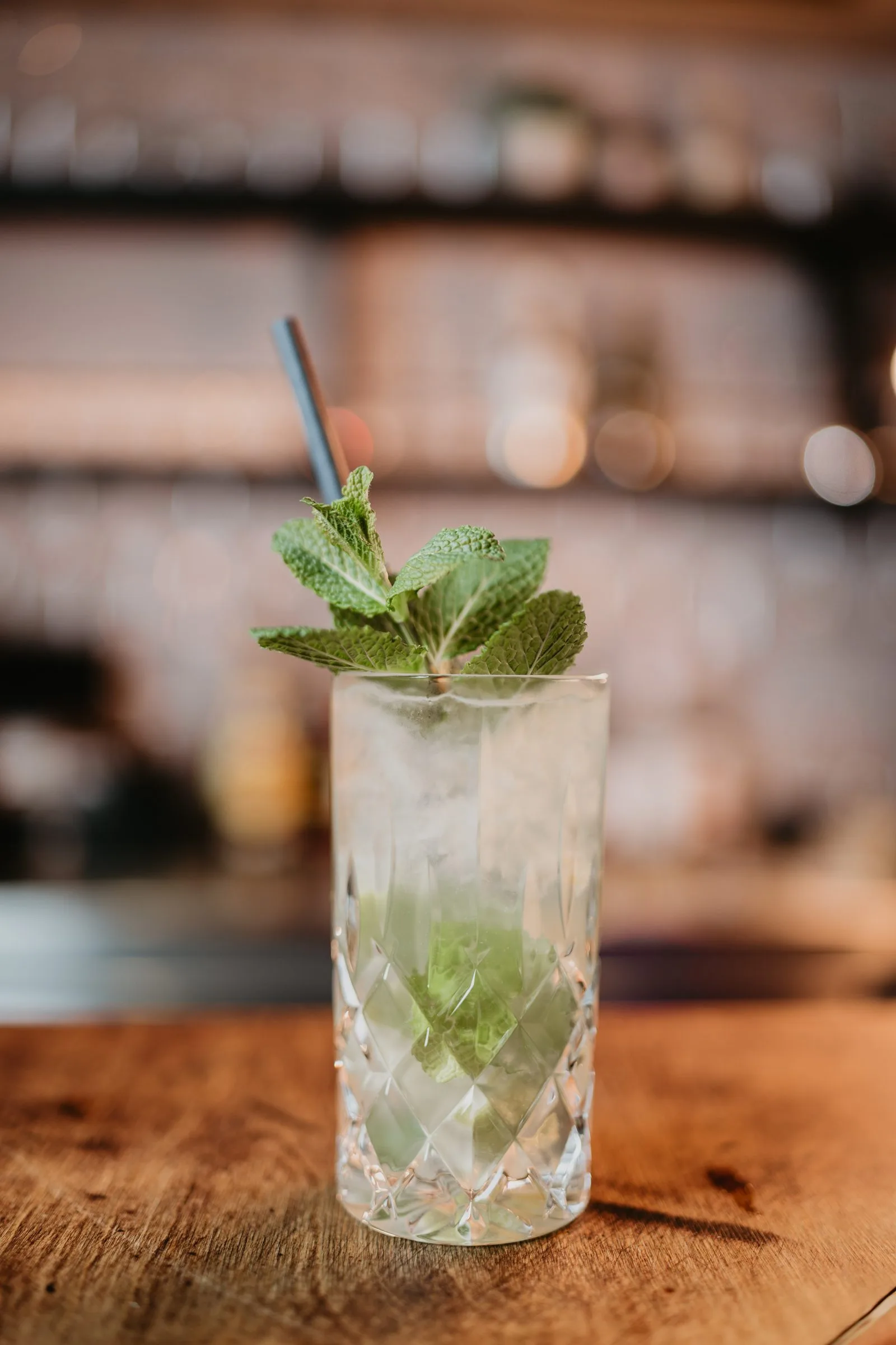 Ein heller Longdrink mit frischer Minze auf dem Tresen, dahinter die beleuchtete Bar.