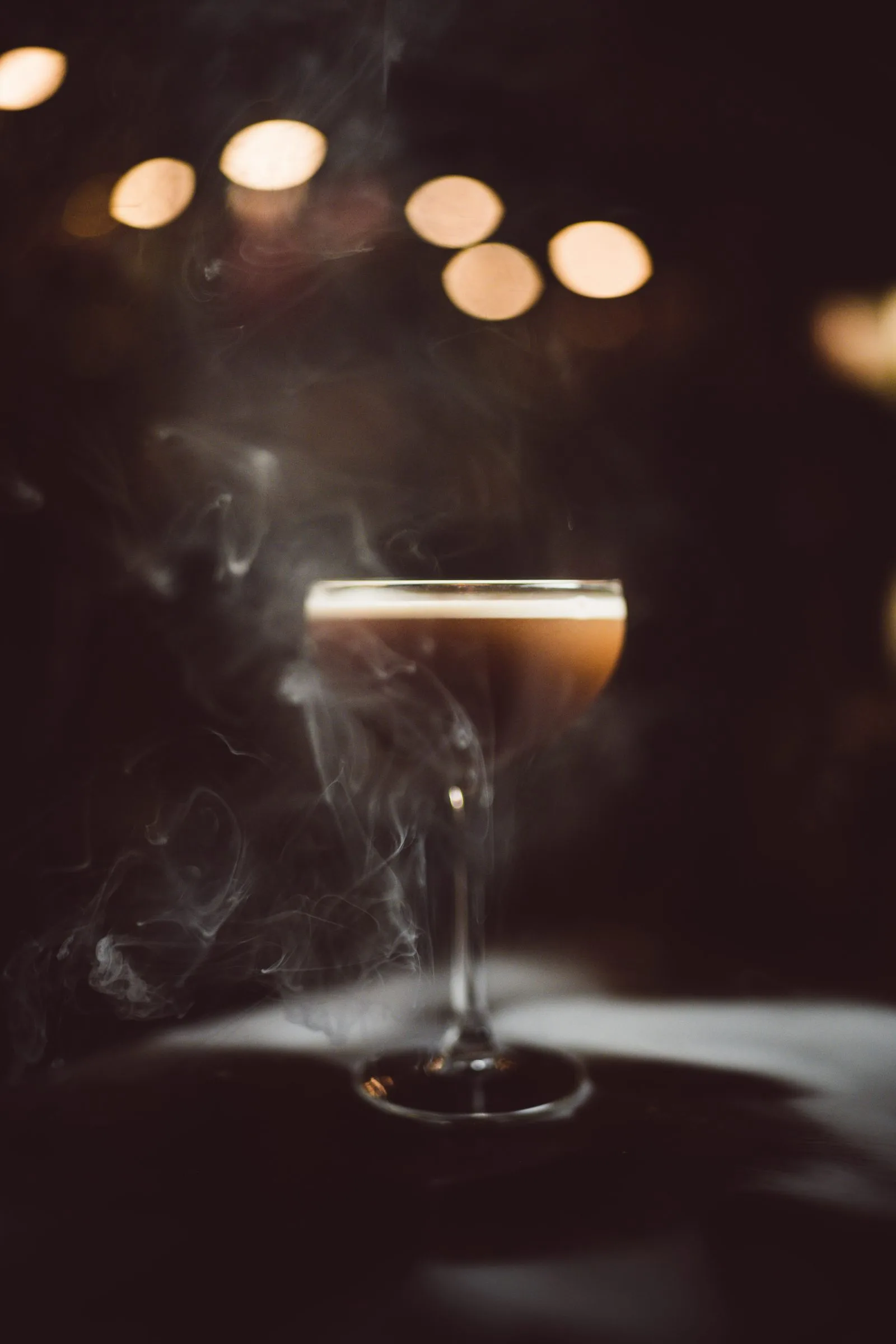 Ein Espresso Martini im Stielglas, umhüllt von Rauch, vor warmen Lichtern.