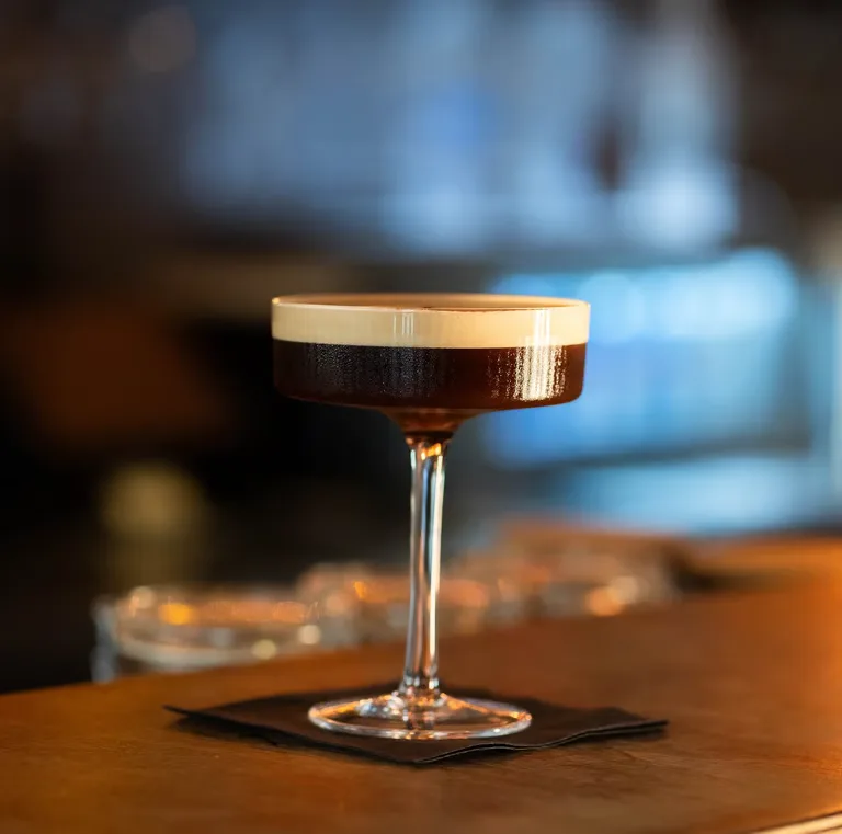 Ein dunkler Cocktail im Stielglas auf dem Holztresen.