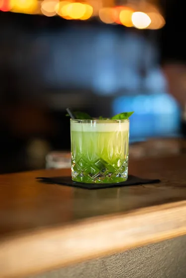 Ein grüner Cocktail mit Kräutergarnitur auf dem Bartresen.