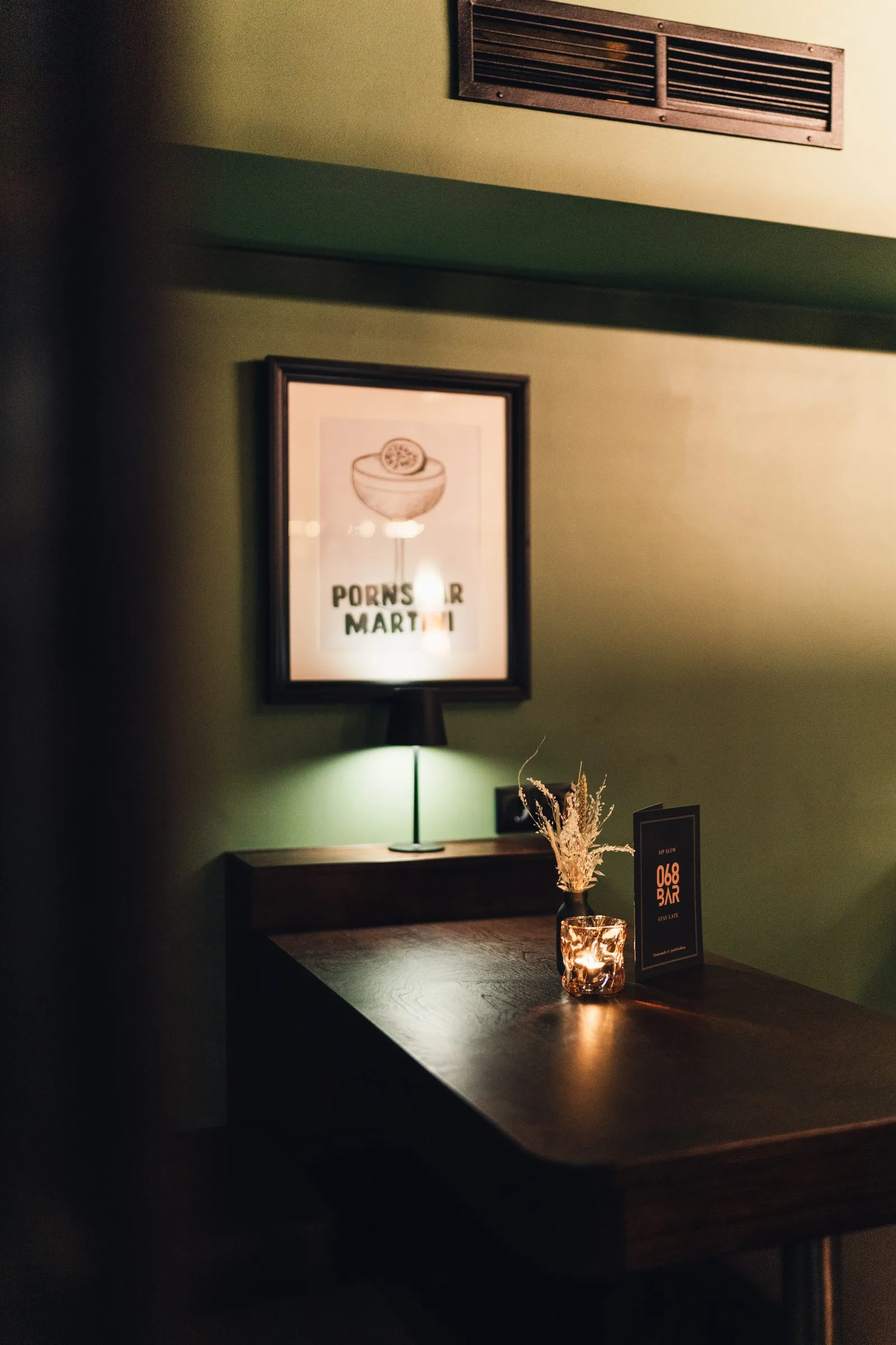 Ein Tisch an der grünen Wand mit Tischlampe, Windlicht und Cocktailposter.