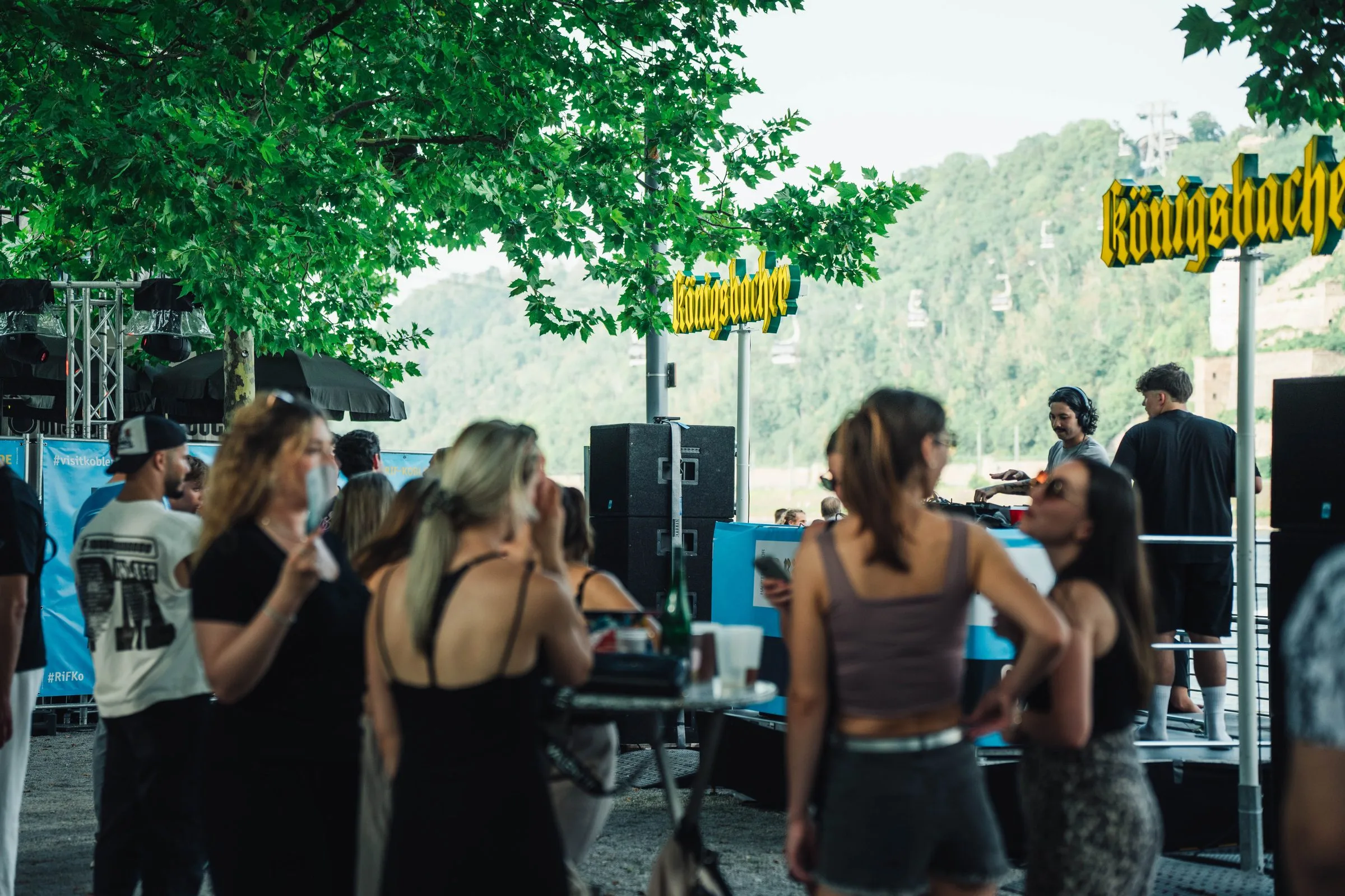 Gäste feiern unter Bäumen am Rheinufer vor dem DJ-Pult.