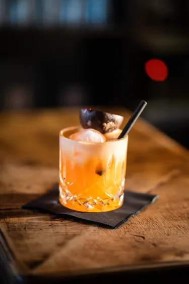 Ein orangefarbener Cocktail mit Eis und Garnitur im Tumbler.